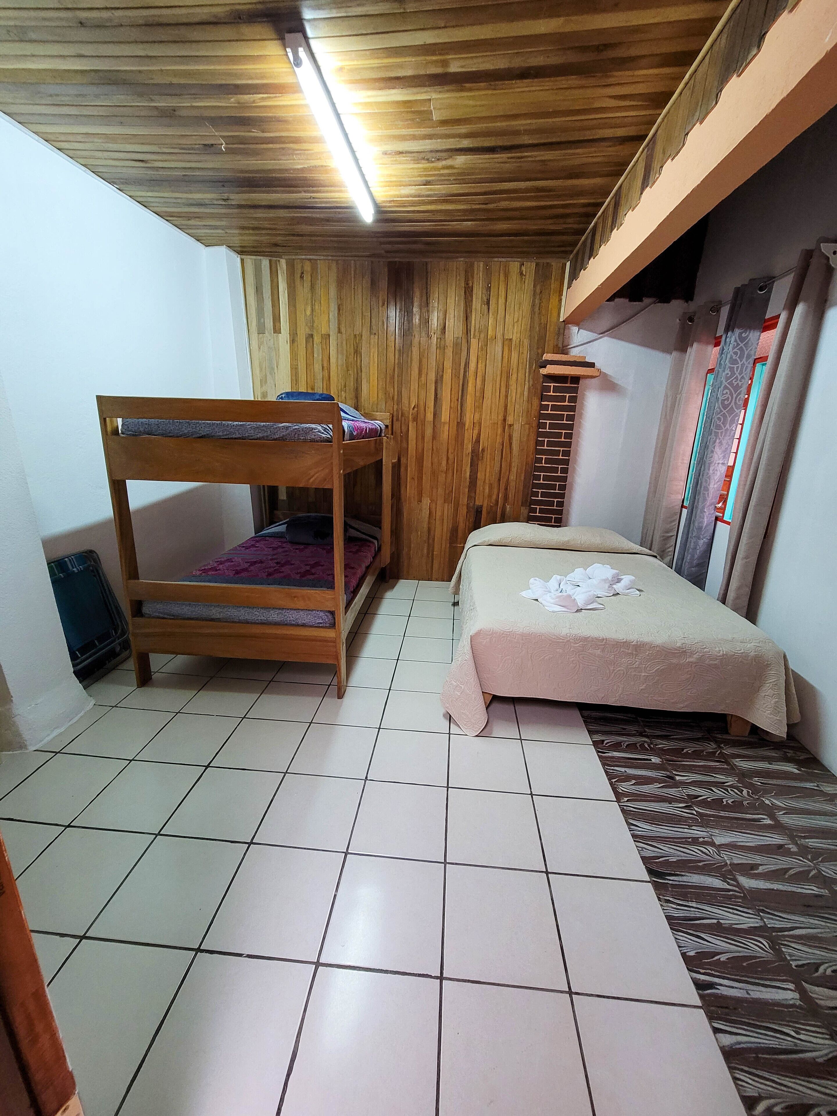 Una habitación con una cama litera, una cama individual, suelo de baldosas, techo de madera y una ventana con cortinas.