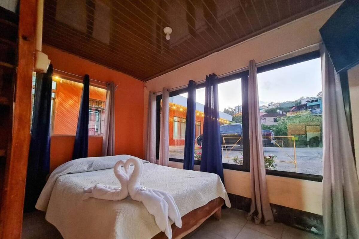 A hotel room with a bed, large windows, curtains, and a view of the outside.