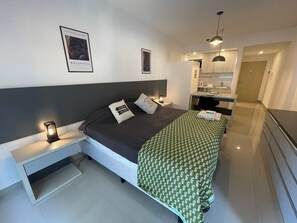 Quarto de hotel moderno com uma cama, mesas de cabeceira, uma escrivaninha e uma cozinha compacta.
