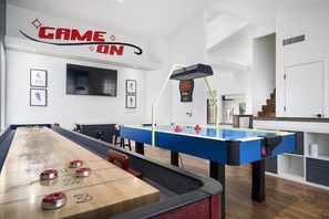A room with a shuffleboard table, a foosball table, and a dartboard. There is a sign that reads "GAME ON" on the wall.