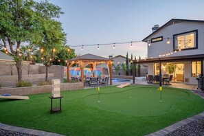 A backyard with artificial grass, a wooden pergola, string lights, and a pool.