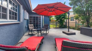 A patio with two lounge chairs, a red umbrella, and a glass door leading to a covered area.