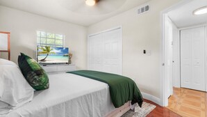 A bedroom with a bed, a television displaying a beach scene, white walls, and a wooden floor.