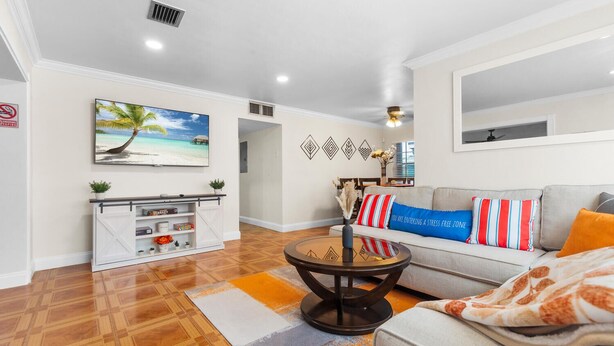 A living room with a sofa, coffee table, and a painting of a beach scene. There is a fireplace with a white cabinet and a plant on top.