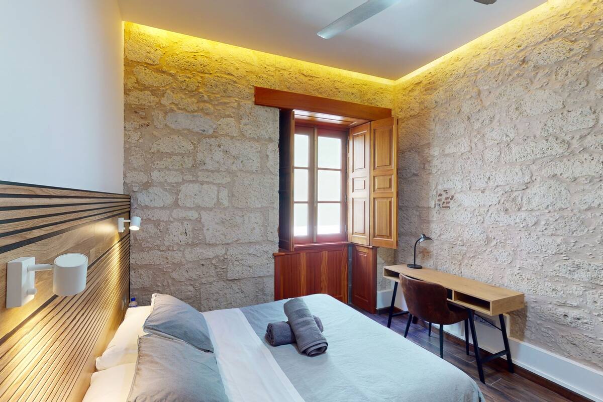 A bedroom with a stone wall, a wooden headboard, a bed with pillows and a rolled towel, a desk with a chair, and a window with wooden shutters.