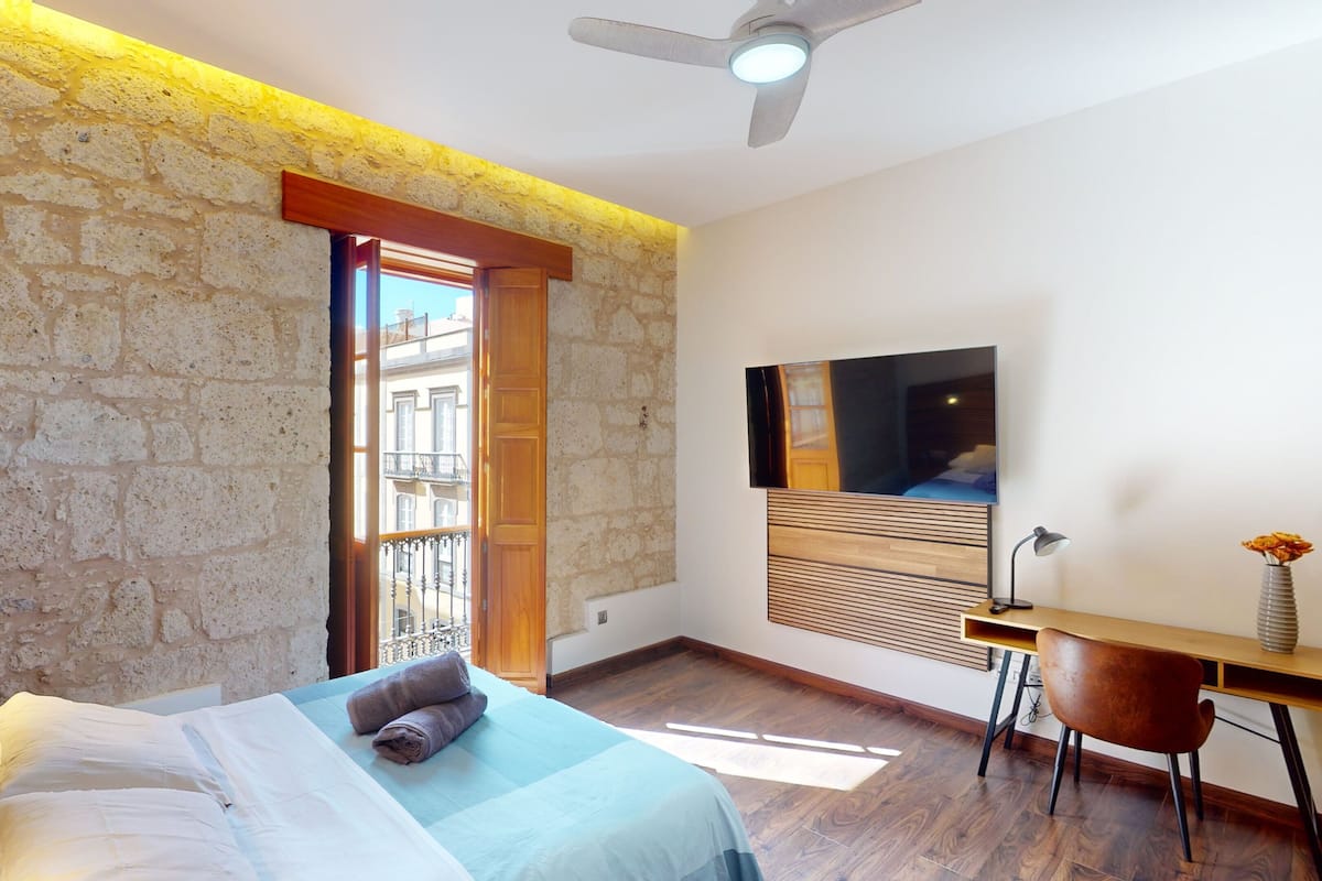 A bedroom with a stone wall, a bed with white and blue bedding, a wooden desk, a chair, a TV, and a ceiling fan.
