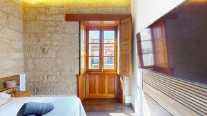 A bedroom with a stone wall, a wooden door leading to a balcony, and a large flat-screen TV mounted on the wall.