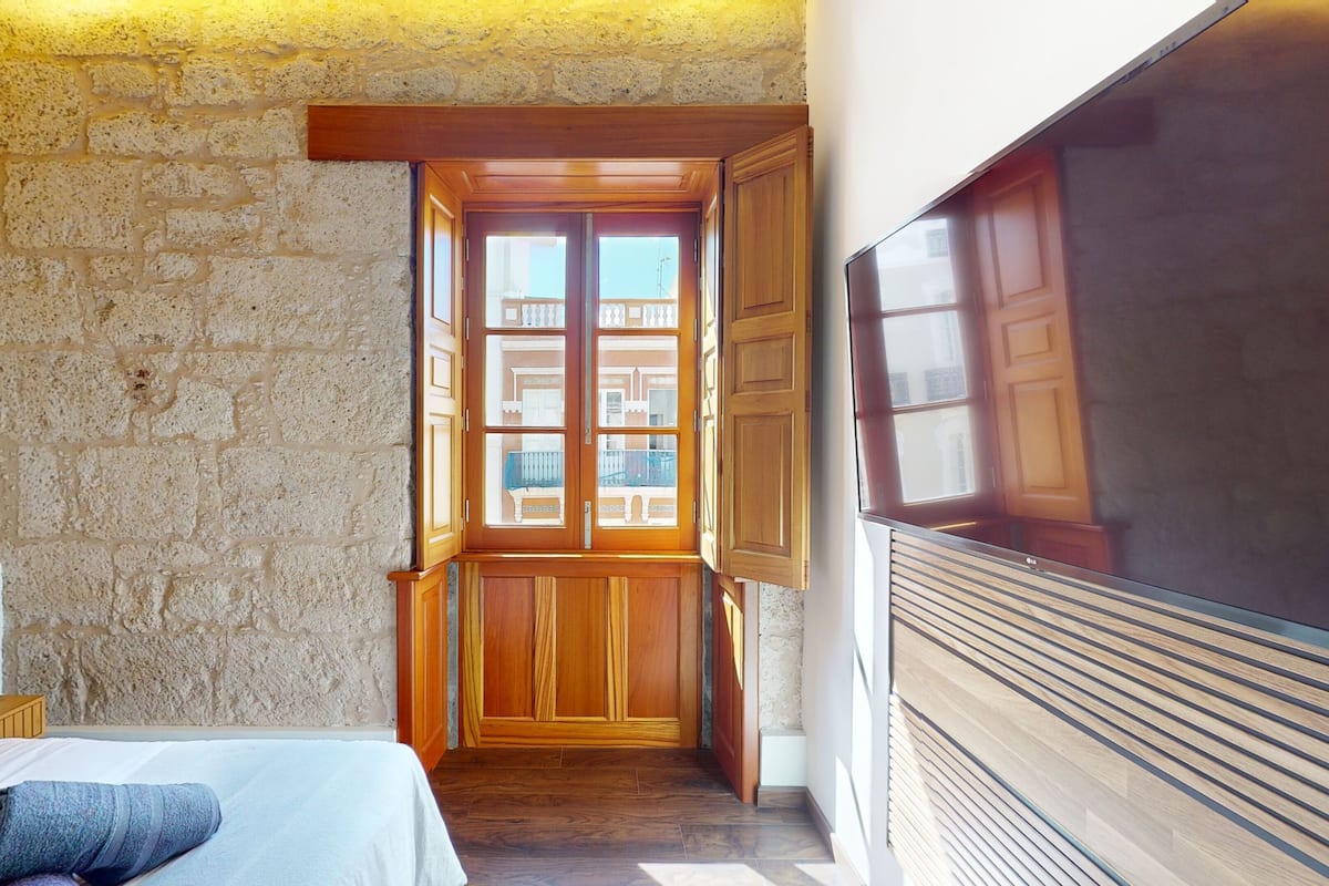 A bedroom with a stone wall, a wooden door leading to a balcony, and a large flat-screen TV mounted on the wall.