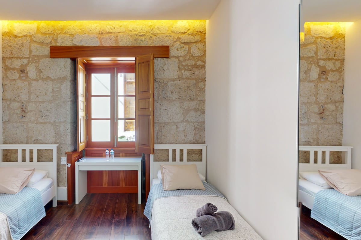 A small, cozy room with two single beds, a wooden desk, and a stone wall.