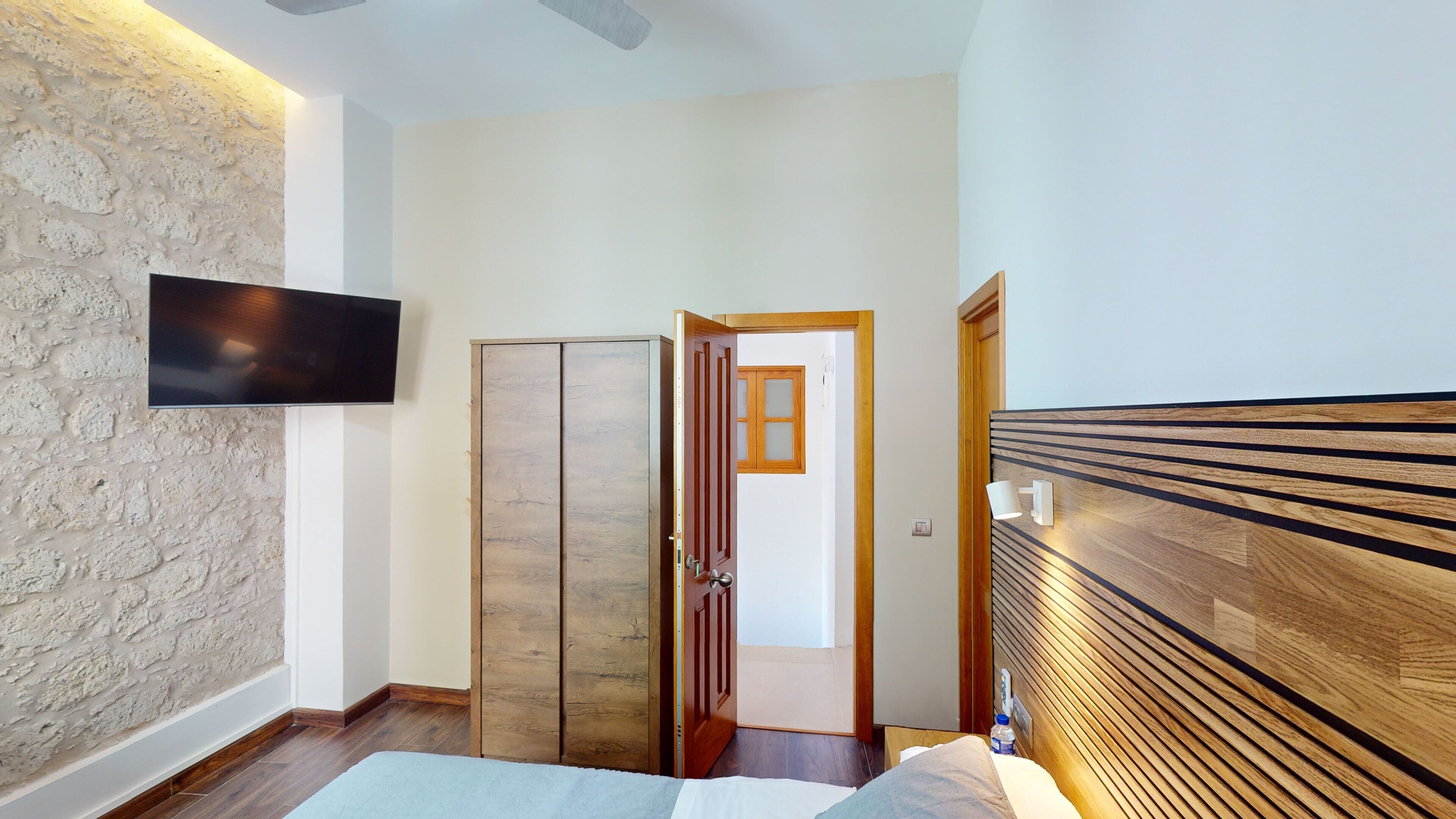 Un pasillo con suelo de madera, un televisor montado en la pared, un armario y una puerta que da a otra habitación.