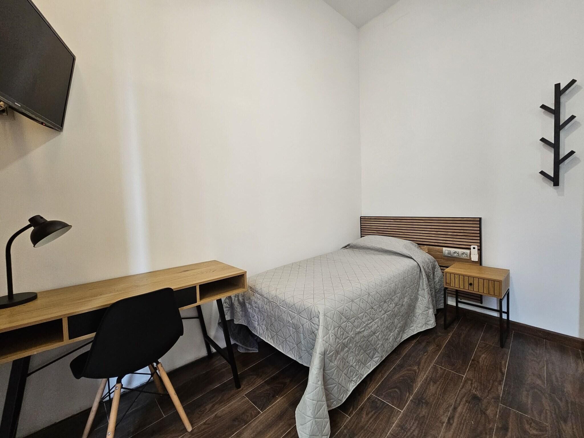 Een slaapkamer met een bed, een bureau, een stoel, een televisie en een wandlamp.