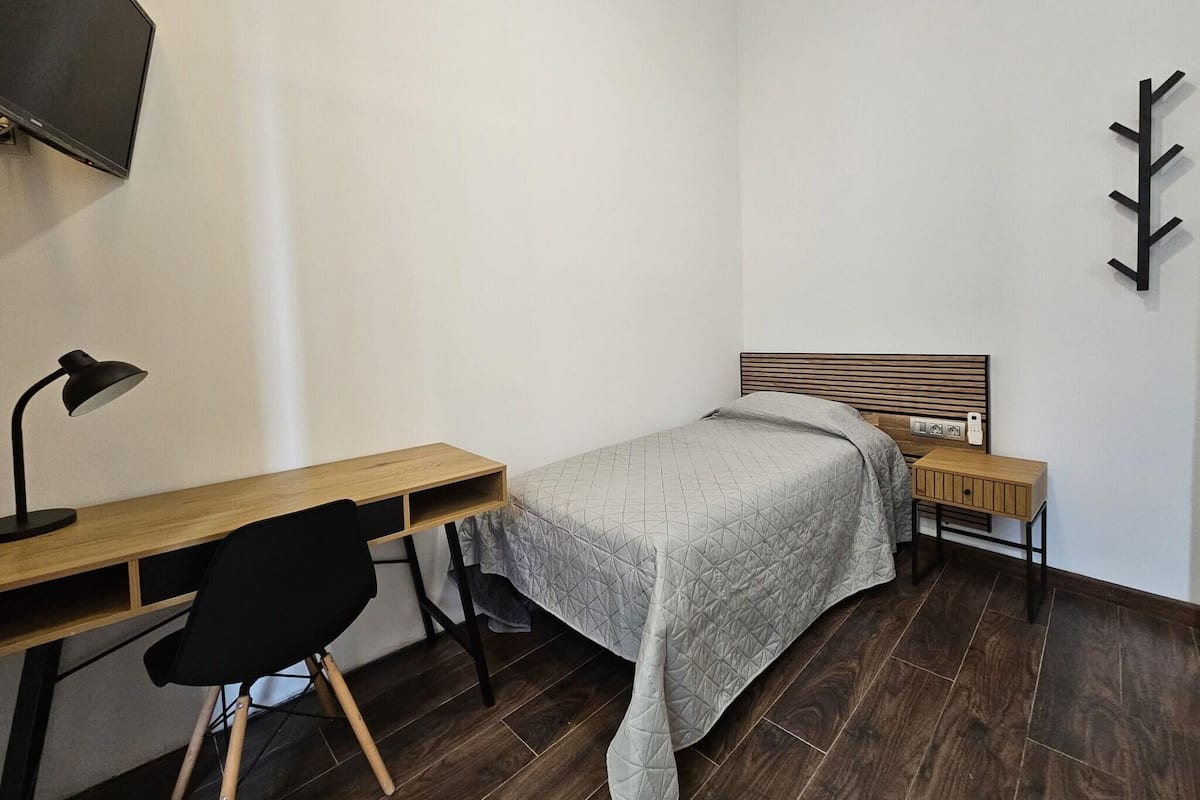 A bedroom with a bed, a desk, a chair, a TV, and a wall-mounted light fixture.