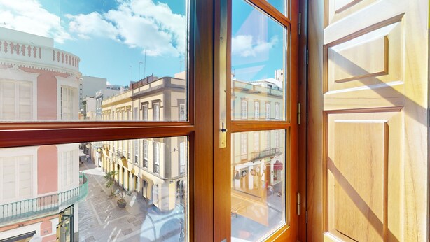 A view from a window with wooden frames, overlooking a street with buildings and a clear sky.