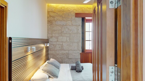 A bedroom with a wooden headboard, a stone wall, a window, and a bed with pillows and a rolled towel.