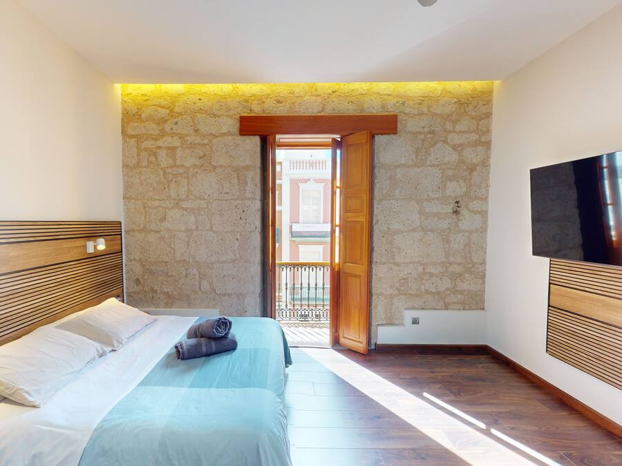 A bedroom with a bed, wooden headboard, stone wall, and a TV mounted on the wall.