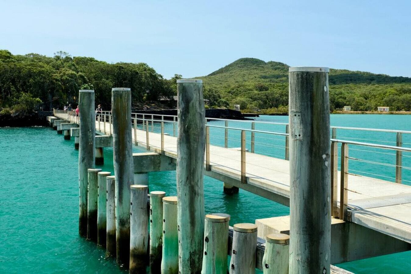 Een houten steiger die zich uitstrekt over turquoise water, met een metalen hekwerk en betonnen pijlers.