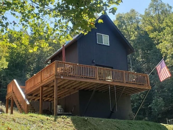 Una cabaña de dos pisos con un balcón y una terraza de madera, rodeada de árboles.