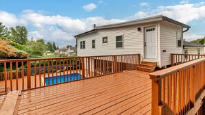 Une terrasse en bois avec une piscine, une maison blanche avec une porte et des fenêtres, et un ciel bleu clair parsemé de nuages.