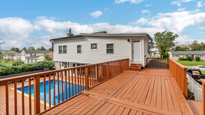 Une terrasse en bois avec une piscine, une maison blanche et un ciel d’un bleu éclatant.