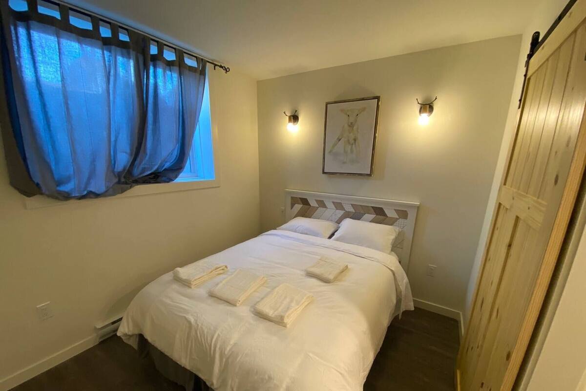 Una cama impecablemente arreglada con sábanas y toallas blancas, una ventana con cortinas azules, un cuadro colgado en la pared y dos apliques.