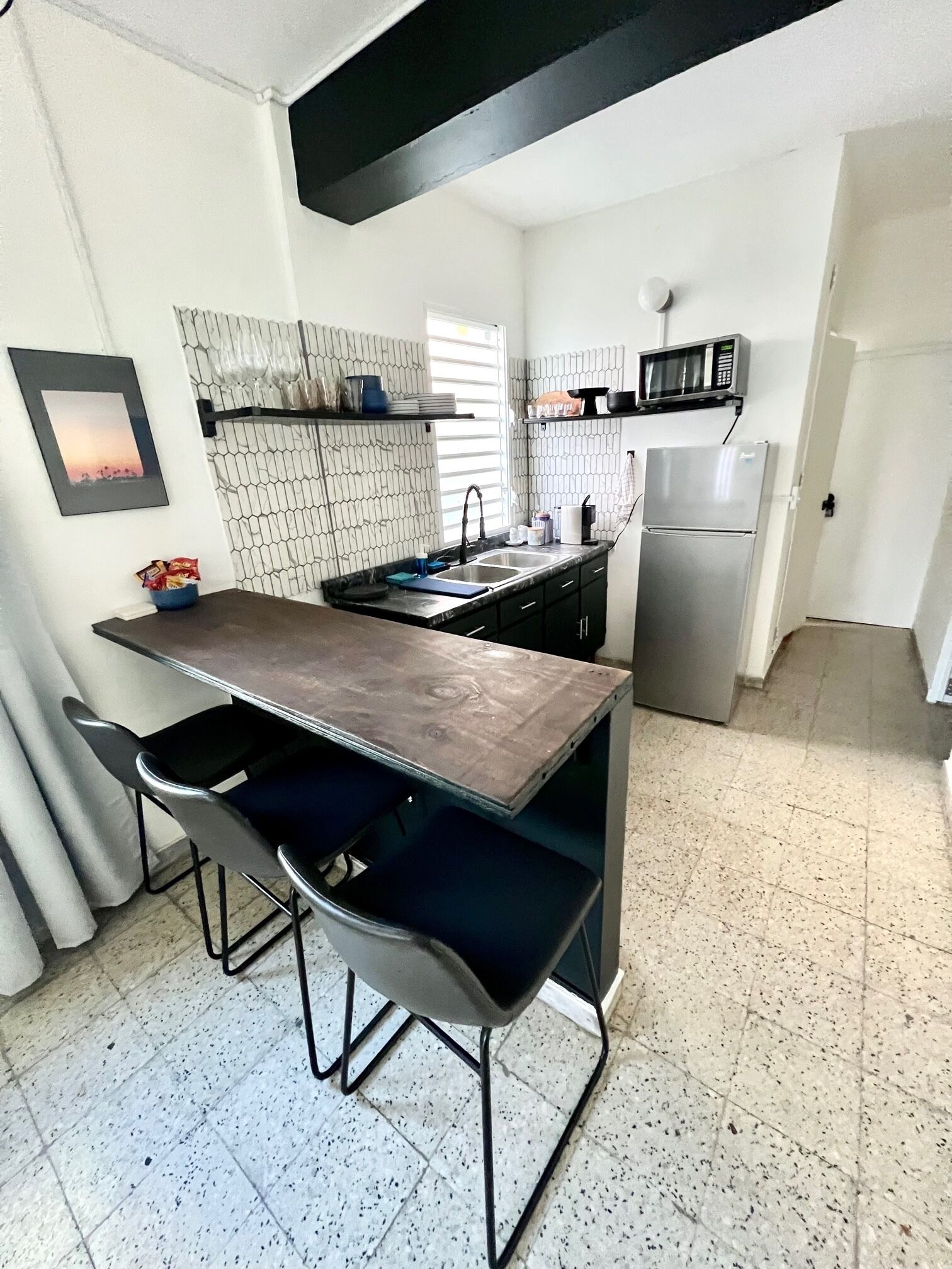 A compact kitchen with a dark countertop, two bar stools, a microwave, and a refrigerator.