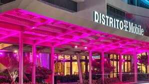 Entrada de un hotel con un área exterior techada, un cartel que dice 'DISTRITO E Mobile' y una fachada bien iluminada.