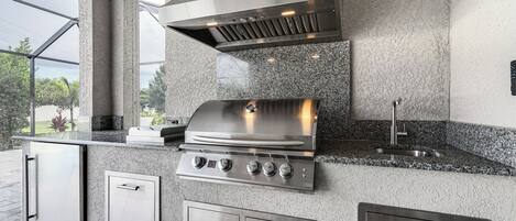 A modern outdoor kitchen with a stainless steel grill, built-in cabinets, and a large window providing a view of the outdoors.