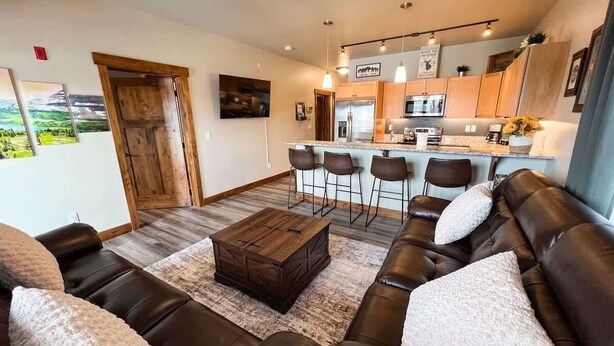 A living room with a brown leather sofa, a wooden coffee table, and a kitchen area with a bar counter and bar stools. - New! Beautiful Condo, Downtown Columbia Falls! (Columbia Falls)