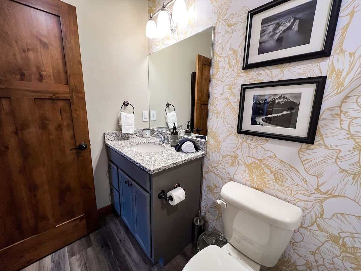 Un baño con puerta de madera, un lavamanos con espejo, un inodoro y cuadros en la pared.