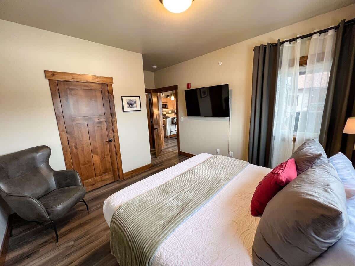 Een slaapkamer met een bed, een stoel, een televisie en een deur die naar een keuken leidt.