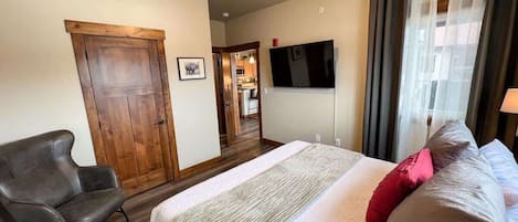 Een slaapkamer met een bed, een stoel, een televisie en een deur die naar een keuken leidt.