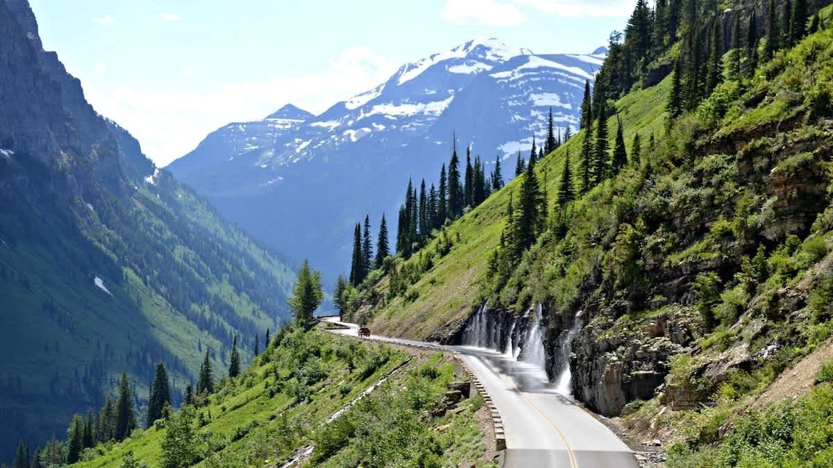 Una carretera de montaña con una cascada a la derecha, rodeada de colinas verdes y montañas nevadas al fondo.