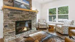 A stone fireplace with a fire, a large window with a view of greenery, and patterned armchairs. - Chill in Style: Luxe Lodge w/ Hot Tub & Rooftop Lounge! (Branson)