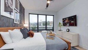 A modern bedroom with a large bed, a ceiling fan, a TV, and a city view.