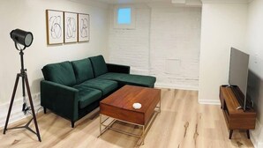 A modern living room with a green sectional sofa, a wooden coffee table, a tripod-mounted camera, a flat-screen TV, and a white brick wall. - 2-Bedroom in the Heart of Denver (Denver)