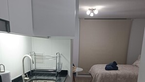 Kompakti hotellihuone, jossa on pieni keittiönurkkaus, kukallinen päiväpeite päällystetty sänky ja pieni pöytä tuolilla.