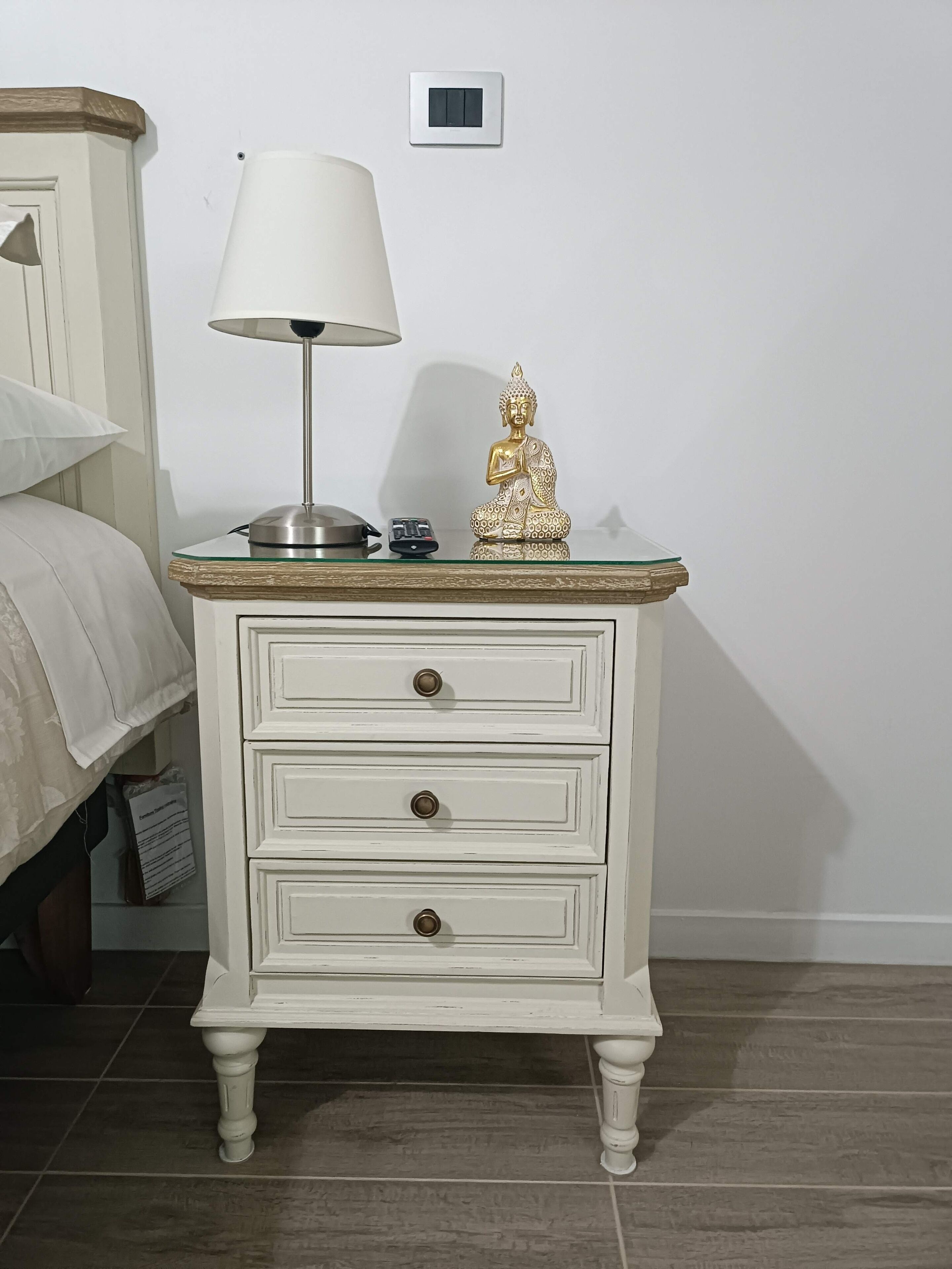 A white bedside table with a lamp, a small Buddha statue, and a remote control.