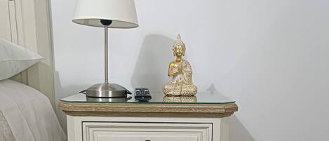 A white bedside table with a lamp, a small Buddha statue, and a remote control.