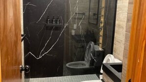 Un baño con piso de mármol, inodoro blanco, lavamanos negro y un espejo.