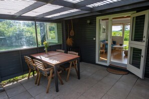 Een overdekt terras met een houten tafel en stoelen, een hangende bezem en een schuifdeur die leidt naar een binnenruimte.