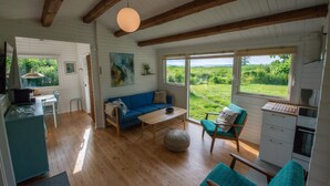A cozy room with a wooden ceiling, a sofa, a coffee table, a dining table, and a kitchen area. - Lovely cottage with wilderness bath and view (Holbæk)