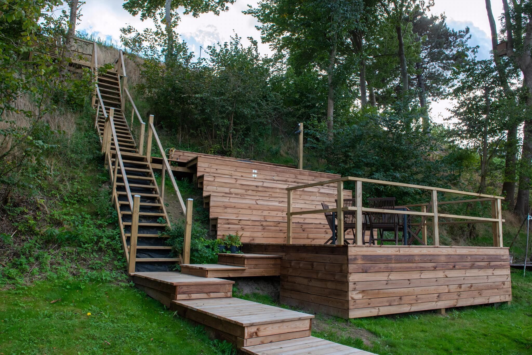 A wooden outdoor structure with a staircase leading up, a seating area with chairs, and a small table.