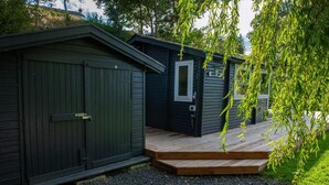A wooden deck leading to a dark-colored building with a door and windows. - Lovely cottage with wilderness bath and view (Holbæk)