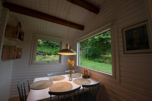 A dining area with a table set for six, surrounded by chairs, a hanging pendant light, and a view of a garden through a window.