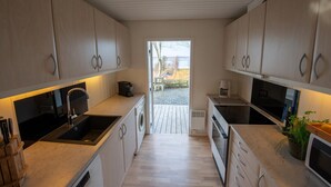 A modern kitchen with wooden cabinets, a built-in oven, a sink, and a window leading to an outdoor area. - Cottage with panoramic view of Lammefjorden (Holbæk)
