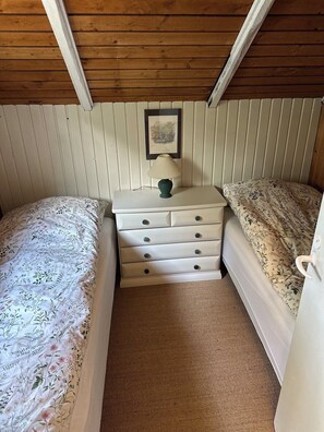 Een knus, klein kamertje met twee bedden, een dressoir, een lamp en een lijst met een afbeelding aan de muur.