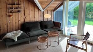 Une pièce chaleureuse avec un plafond en bois, un canapé, deux petites tables d’appoint en osier, un fauteuil et une grande fenêtre.