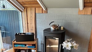 Une pièce avec un poêle à bois noir, une étagère en bois sur laquelle sont posés divers objets, un sac noir et un vase avec des fleurs blanches.