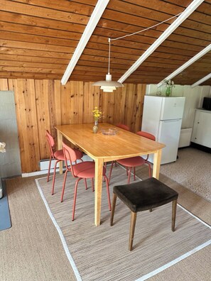 Een houten eettafel met stoelen, een klein krukje, een hanglamp en een koelkast in een kamer met houten wanden en een schuine kap.