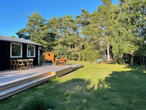 A wooden deck with outdoor furniture, a small building with windows, and a forested area in the background. - Summer house with outdoor sauna at Sjællands Odde (Sjællands Odde)